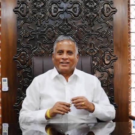V. Somanna seated at an office desk in front of a carved wooden panel, smiling