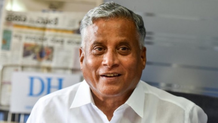 V. Somanna smiling during a conversation, wearing a white shirt, with newspapers in the background