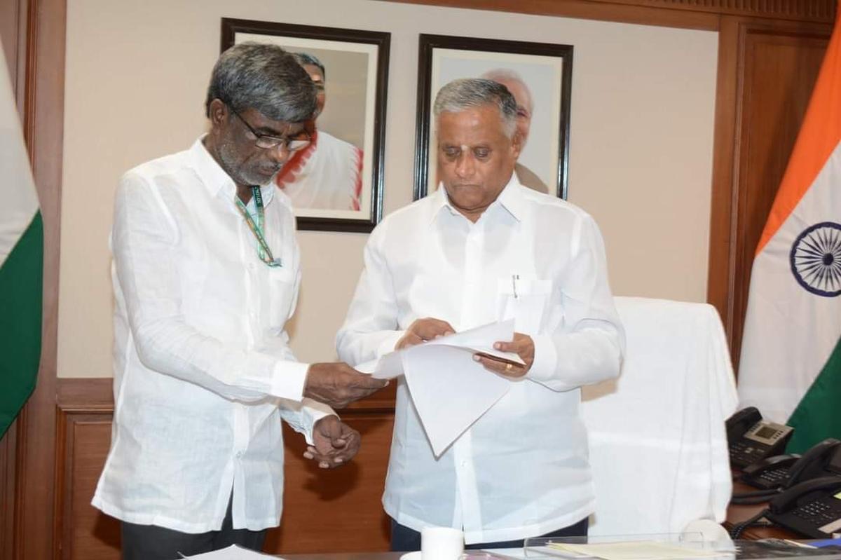 V. Somanna reading a document handed to him by a colleague in an office with the national flag and framed portraits
