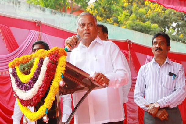 V. Somanna speaking into a microphone at an outdoor public programme, with a floral garland on the lectern and trees behind