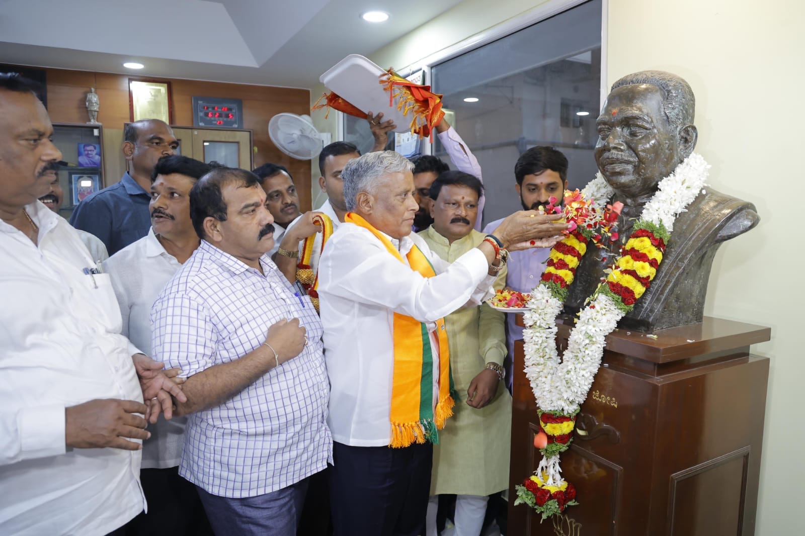 V. Somanna, wearing a saffron and green shawl, offering a floral tribute at a bronze bust garlanded with flowers, surrounded by people