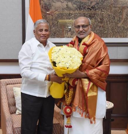 V. Somanna presenting a bouquet of white flowers to a man in a silk shawl, with the national flag behind