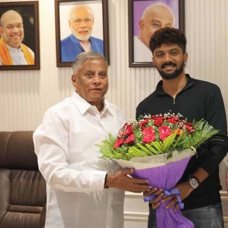 V. Somanna receiving a bouquet of red roses from a young man in an office with framed portraits on the wall