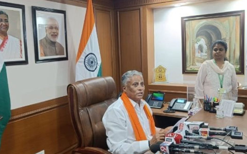 V. Somanna, wearing a saffron shawl, seated in his office chair addressing media microphones, with the national flag and framed portraits on the wall