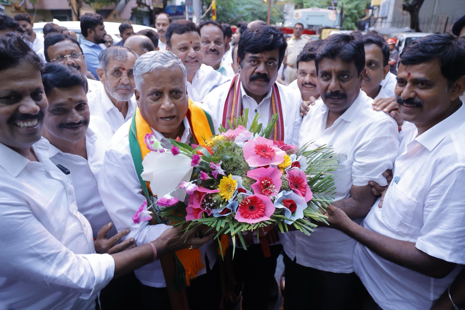 V. Somanna, wearing a saffron shawl, receiving a bouquet from a group of people outdoors