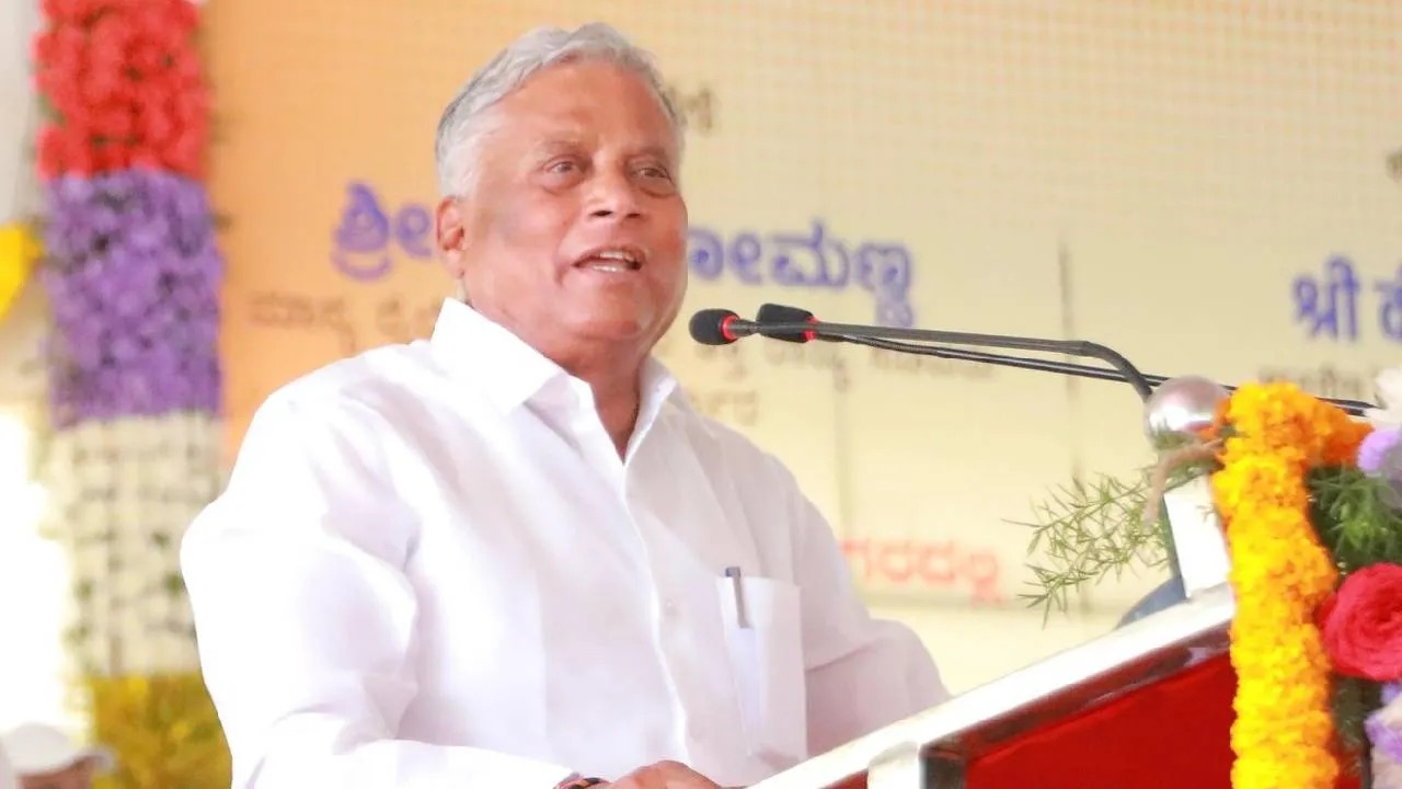 V. Somanna speaking at a lectern at a public programme, with a Kannada-language banner and flower decorations behind him