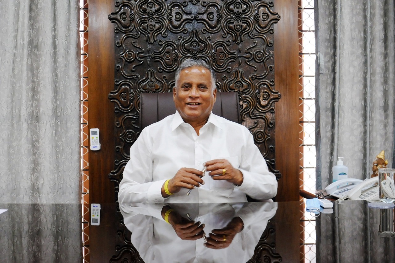 V. Somanna smiling, seated at a large desk with a reflective surface, in front of a carved wooden panel