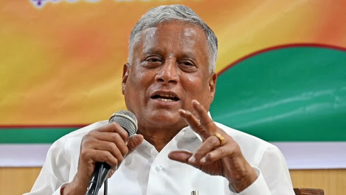 V. Somanna speaking into a microphone and gesturing with his right hand, against a green and orange backdrop