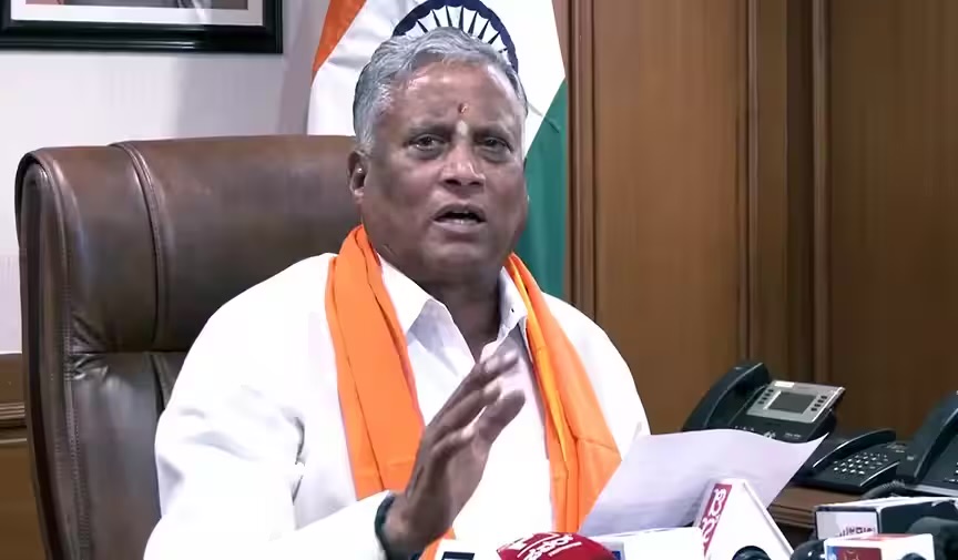 V. Somanna, wearing a saffron shawl, addressing the media from his office chair with microphones on the desk