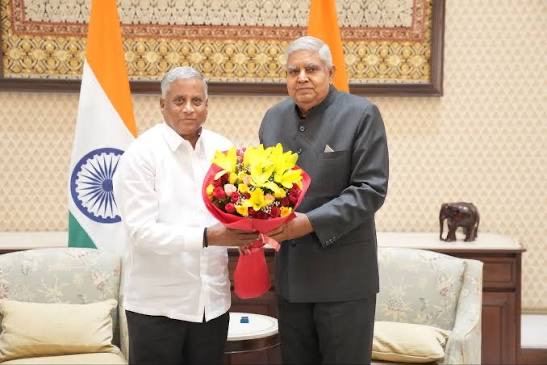 V. Somanna presenting a bouquet of yellow flowers during a formal courtesy call, beside the national flag