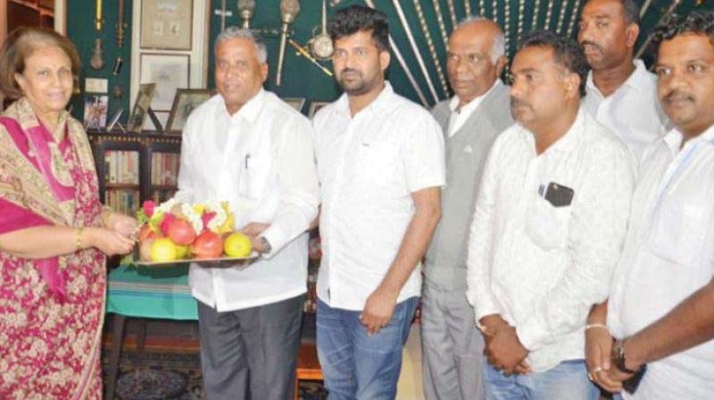 V. Somanna holding a tray of fruit during a courtesy visit, with several people standing beside him in a room with bookshelves