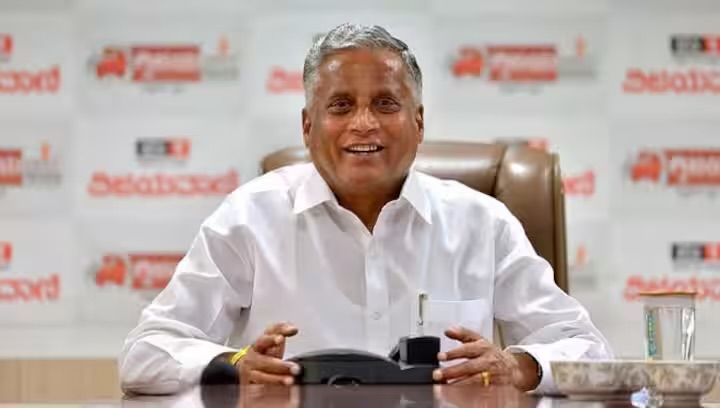 V. Somanna seated at a desk, smiling, in front of a backdrop printed with Kannada media logos