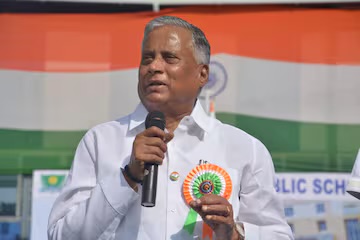 V. Somanna speaking into a microphone at an outdoor event, wearing a tricolour rosette, with a large national flag behind him