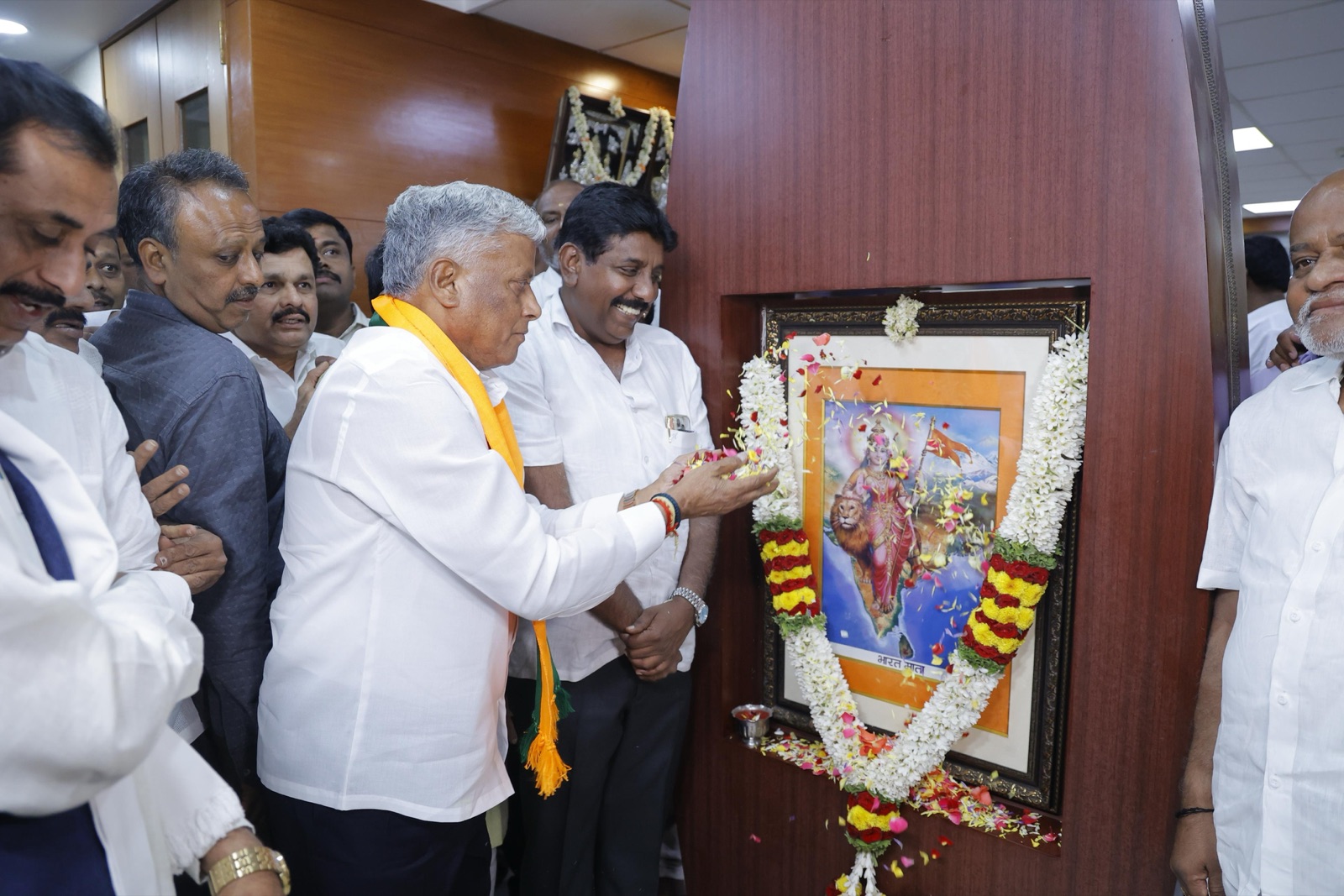 V. Somanna, wearing a yellow shawl, placing flowers before a garlanded framed portrait, surrounded by several people